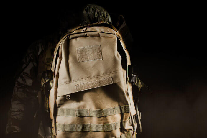 Photo of a fully equipped soldier in uniform, armor and mask holding backpack on dark background with fog and dust close-up view.