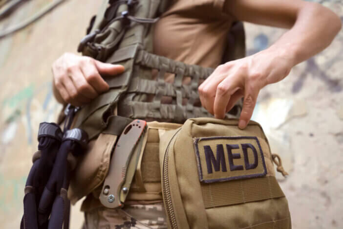 A doctor in a bulletproof vest, in camouflage, opens a first aid kit, a woman is engaged in tactical medicine and saves lives on the front line, a close-up hand.