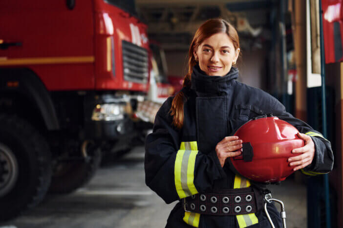 Holds red hat in hands. Female firefighter in protective uniform standing near truck.