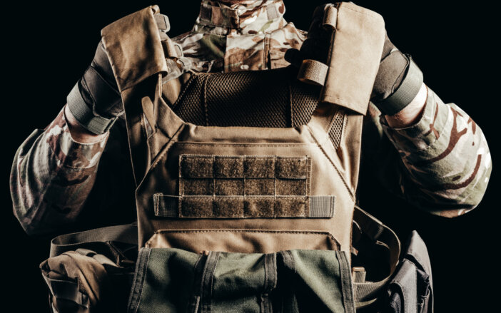 Photo of soldier in camouflaged uniform and tactical gloves holding military armored vest on black background.