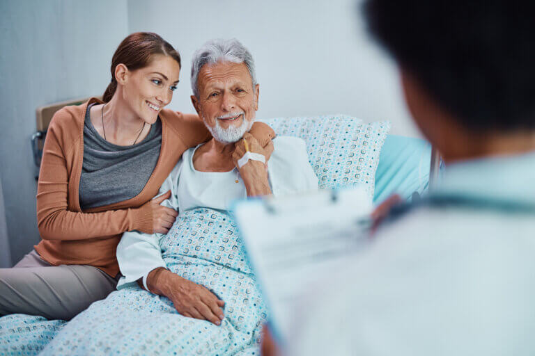 Happy woman embracing he mature father who is recovering at hospital ward and talking to a doctor.