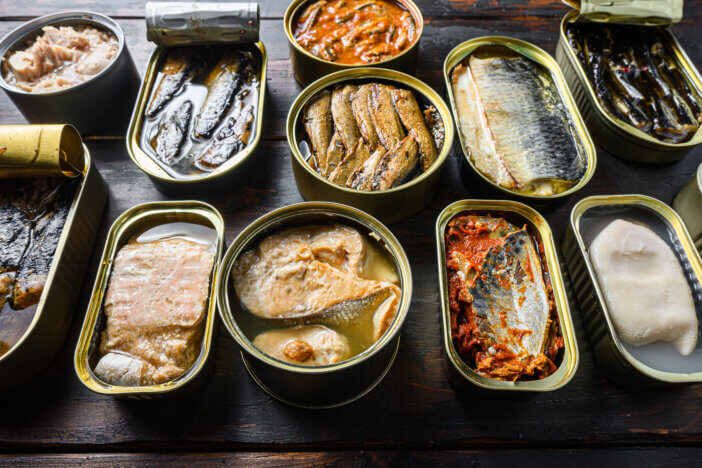 Opened preserve cans with Saury, mackerel, sprats, sardines, pilchard, squid, tuna over vintage homemade wood table close up side view.