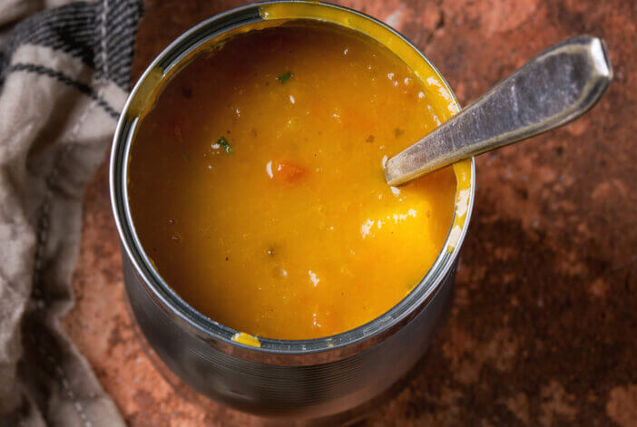 Tin can of carrot soup with spoon, standing on kitchen towel and clay board over old rusty iron background. Dark rustic style. Top view