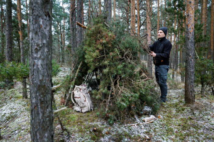 wilderness survival camp - man building tree branch hut in forest in winter