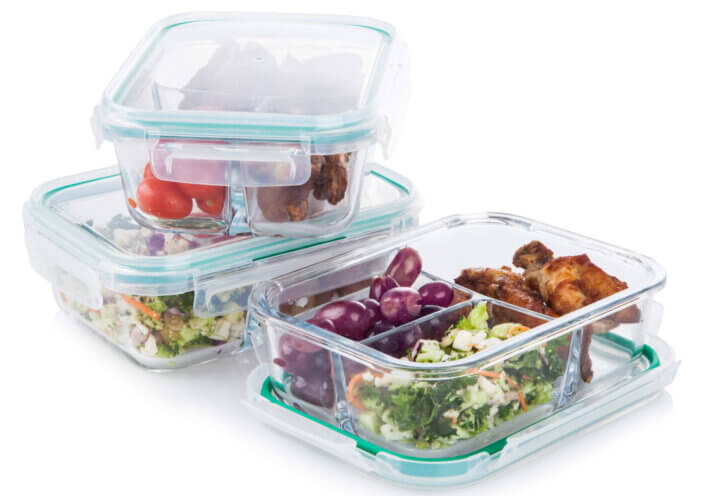 Clear glass food storage meal prepping containers with fresh berries, carrots, fruits, vegetables and meat on white background.