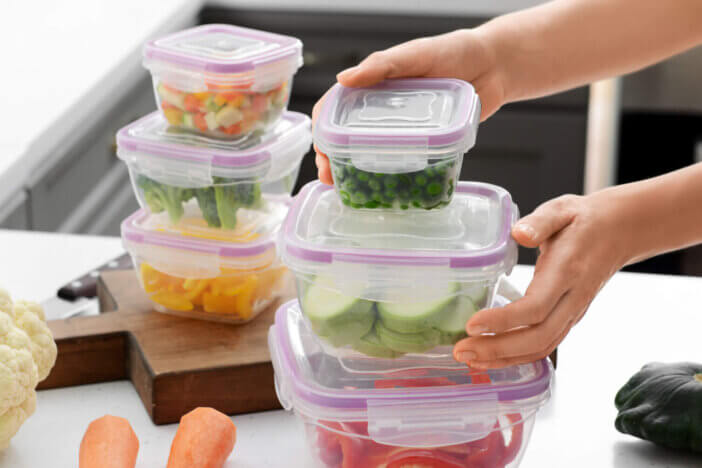 Woman holding stack of plastic containers with fresh vegetables for freezing at table in kitchen