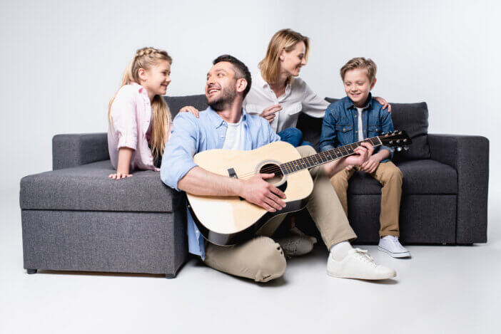 smiling man playing acoustic guitar with family sitting on sofa near by