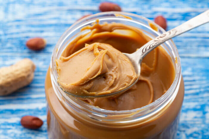 Peanut butter in an open jar and peanuts in the skin are scattered on the blue table. Space for text. Closeup.