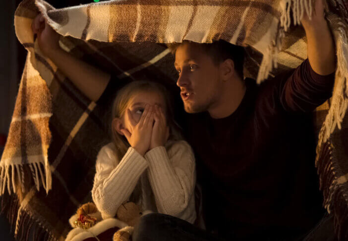 Father telling horror Christmas story for little girl sitting under cozy plaid