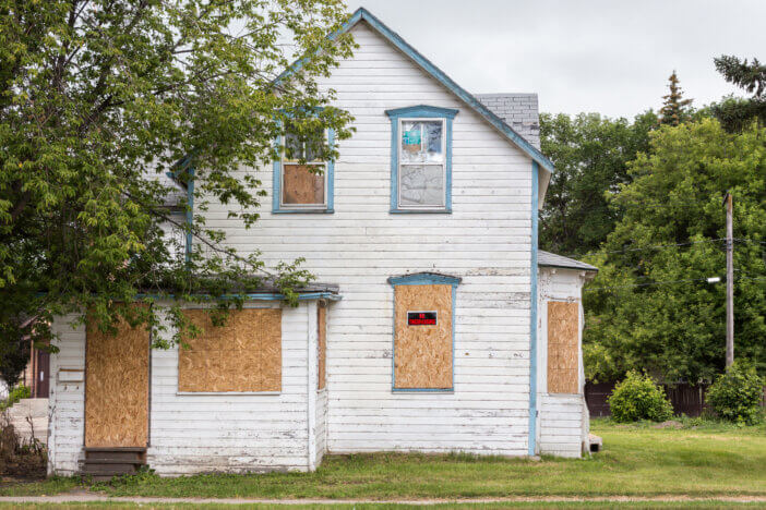an old white condemned two-story house with windows boarded up and a sign saying "no trespassing" with a big green tree in front.