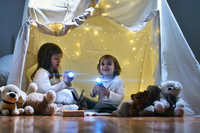 Two little girls play at home in the evening to build a camping tent to read books with a flashlight and sleep inside. Concept of: game, magic, creativity, alarm systems