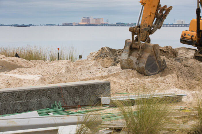 A backhoe at a coastal resiliency project during construction of a seawall on the shores of the Bay of Biloxi in Ocean Springs, Mississippi with a Biloxi Casino and a river barge in the background.