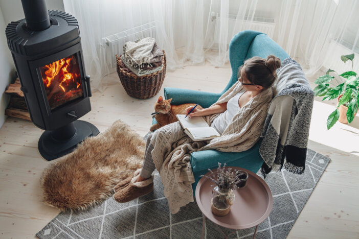 Young woman write in a notebook sitting in a cozy armchair by the fireplace with a domestic cat
