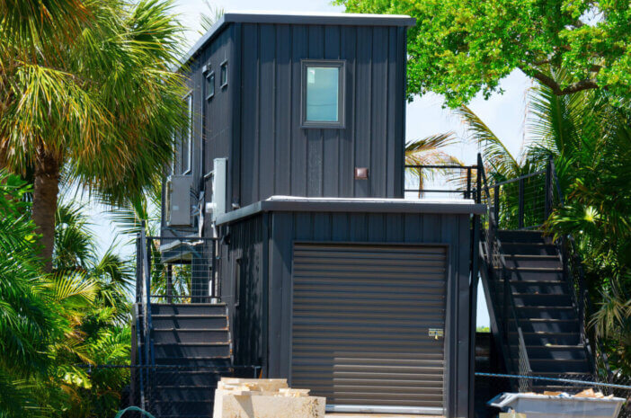Metal shipping container house under construction with building material products in front yard on piece of land with lush green trees.