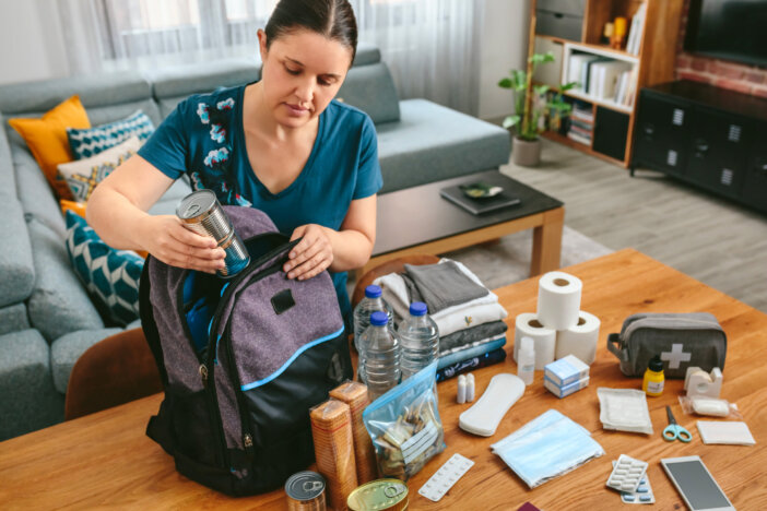 Woman putting cans of food to prepare emergency backpack in living room